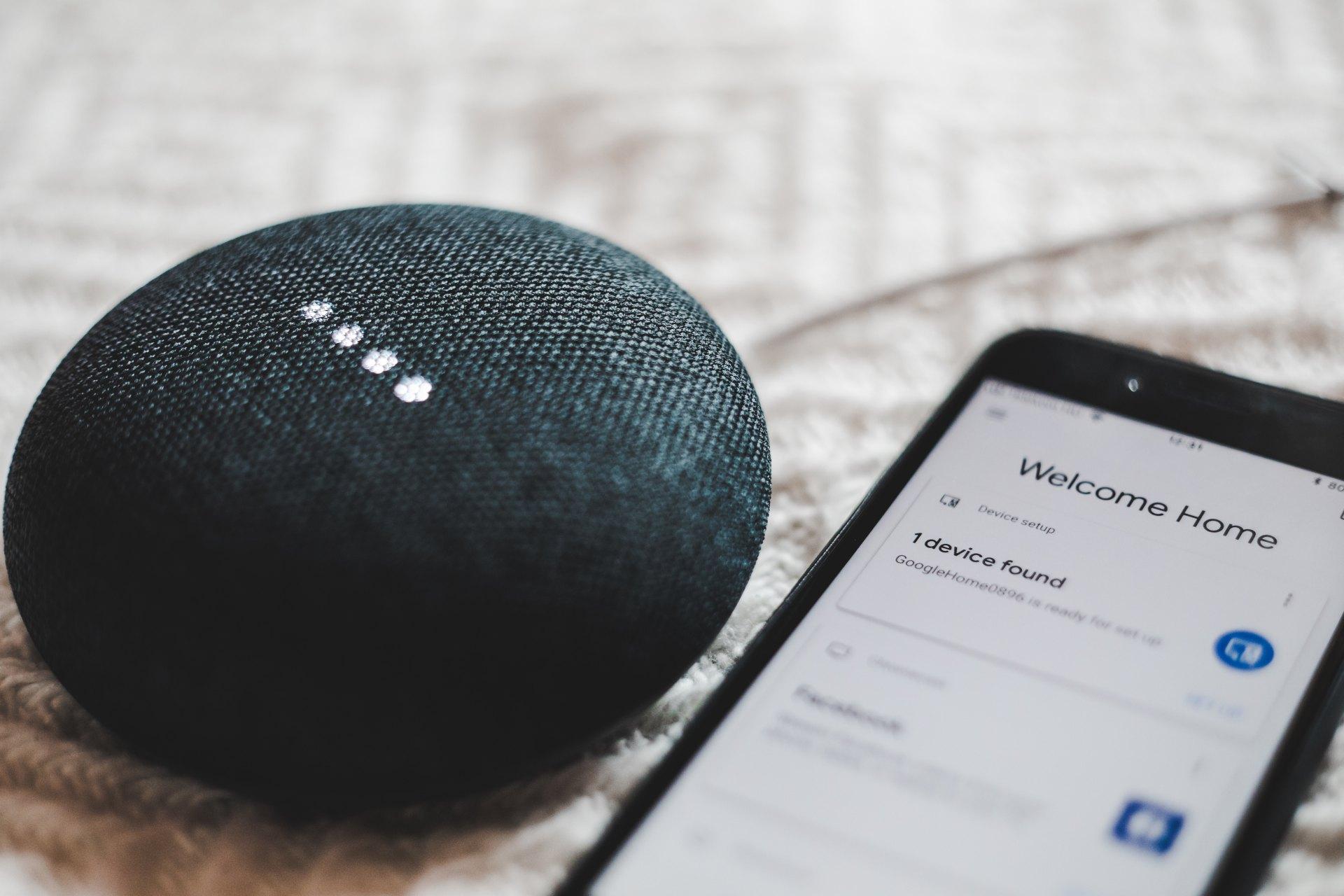 Smart speaker beside a smartphone showing a home setup screen