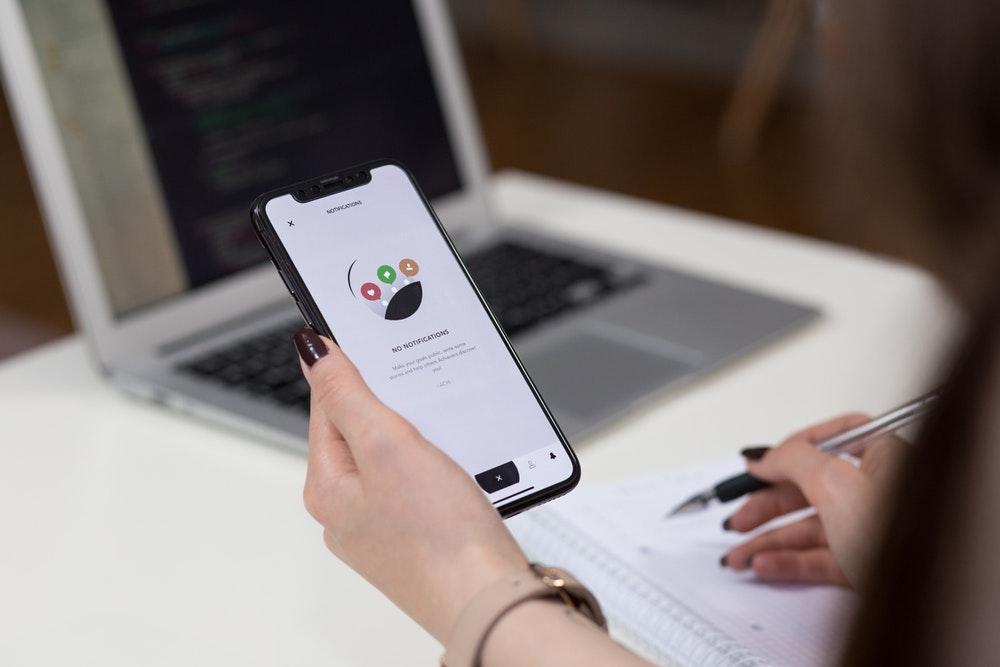 Person holding a smartphone beside a laptop and notebook