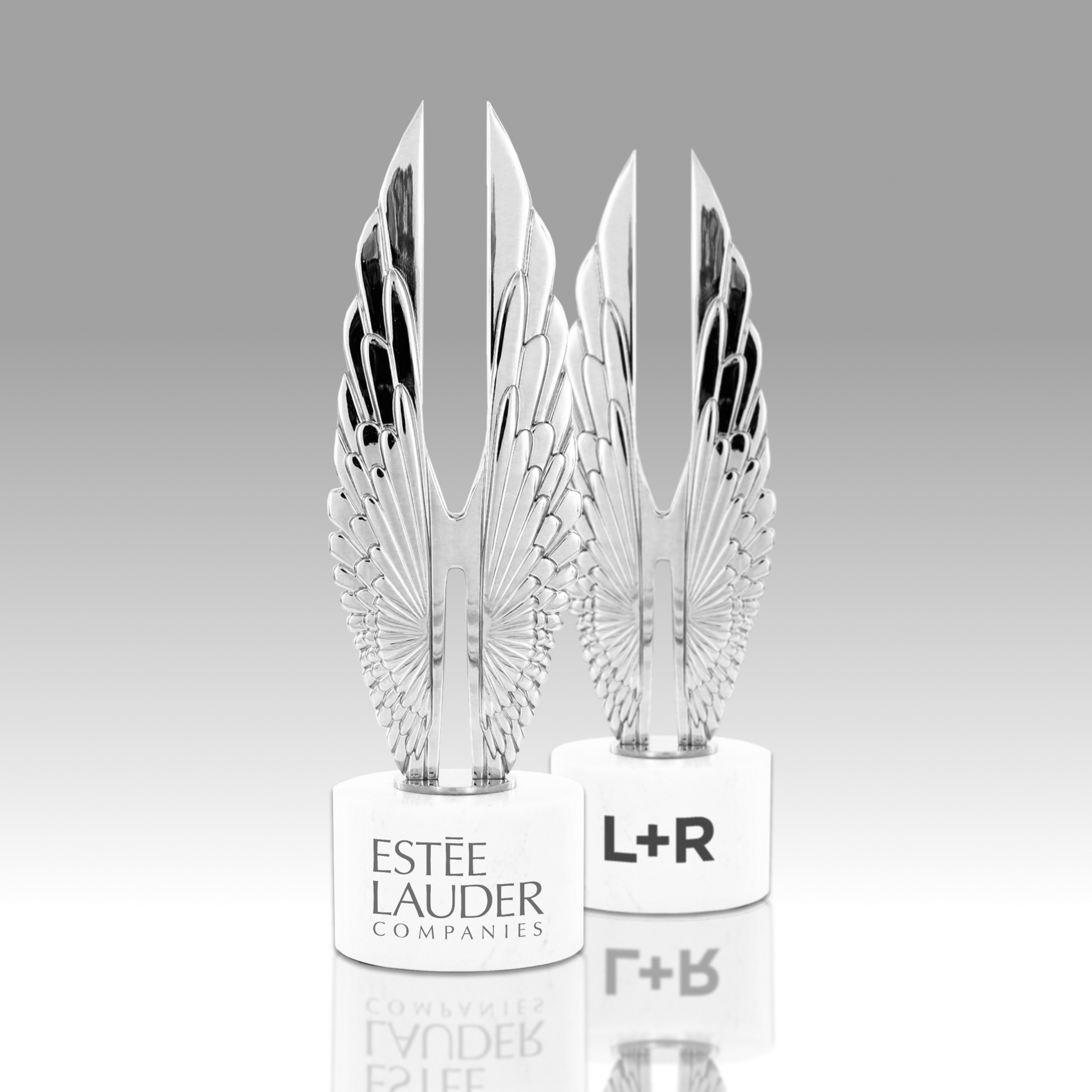 Two silver wing-shaped trophies on white bases labeled Este Lauder and L+R