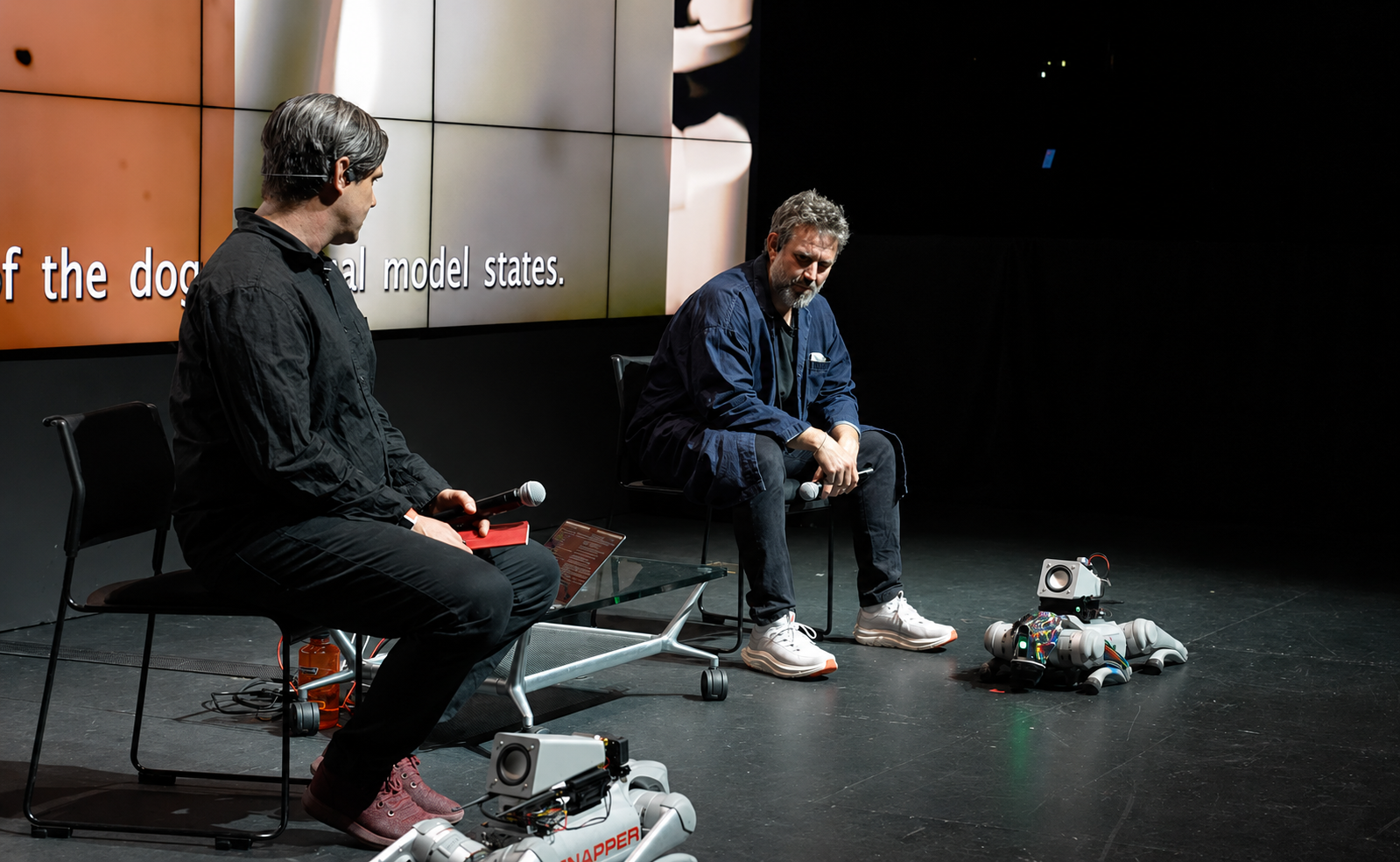 Two speakers sit onstage beside small companion robots.
