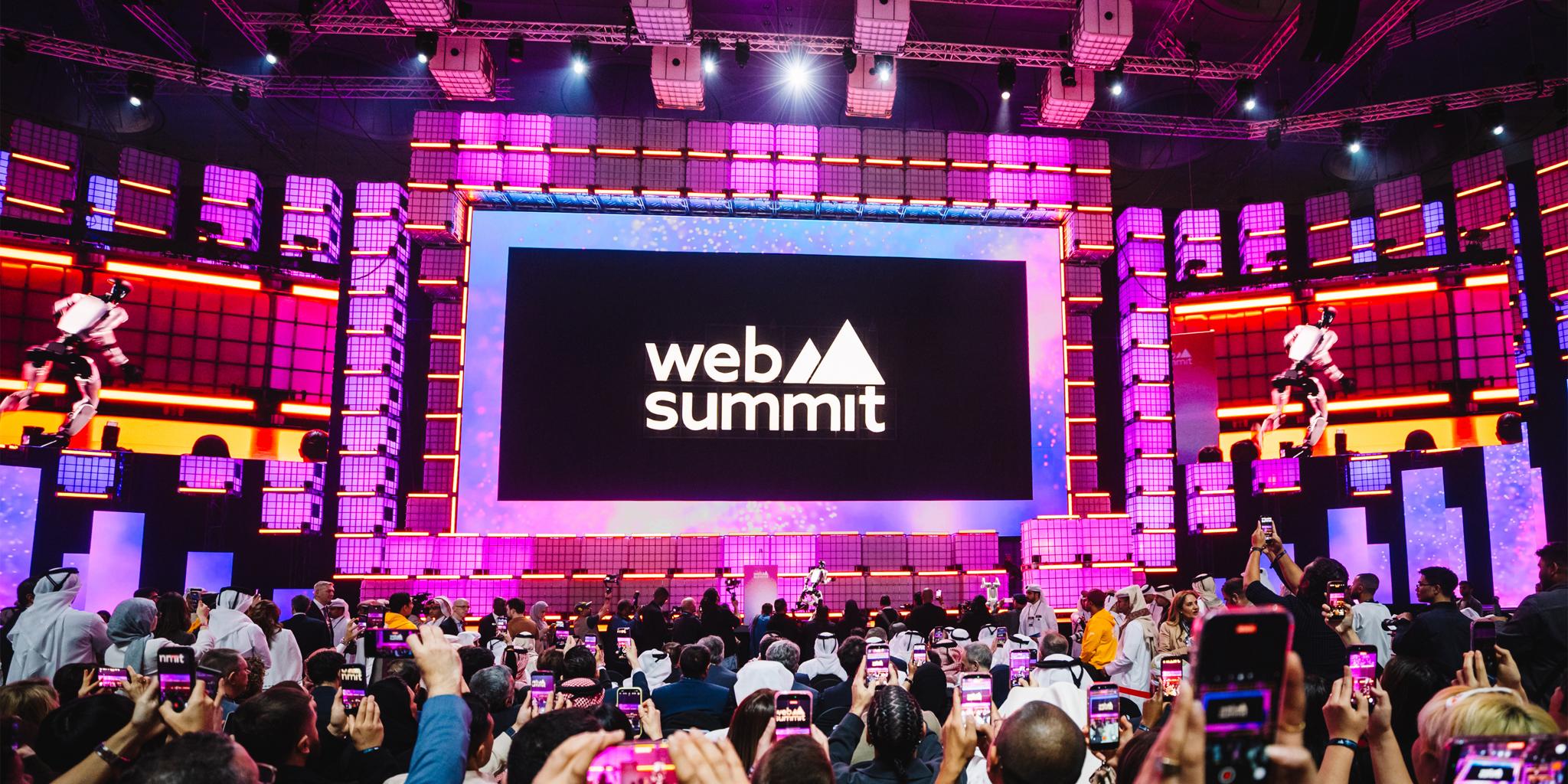 Crowd recording a brightly lit Web Summit stage with large screen and pink lighting.