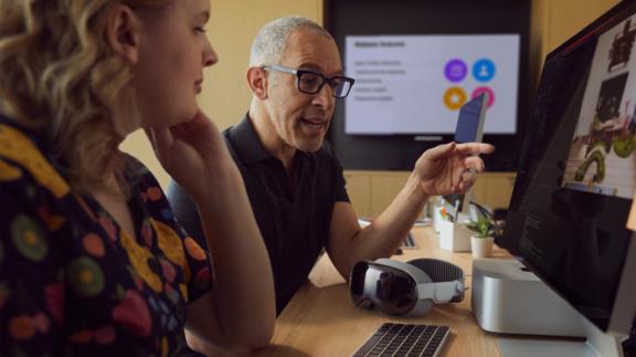 Two coworkers review a desktop screen with a mixed reality headset on the desk