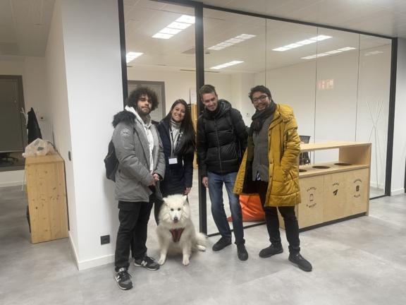 Four people pose with a white dog in a modern office hallway.