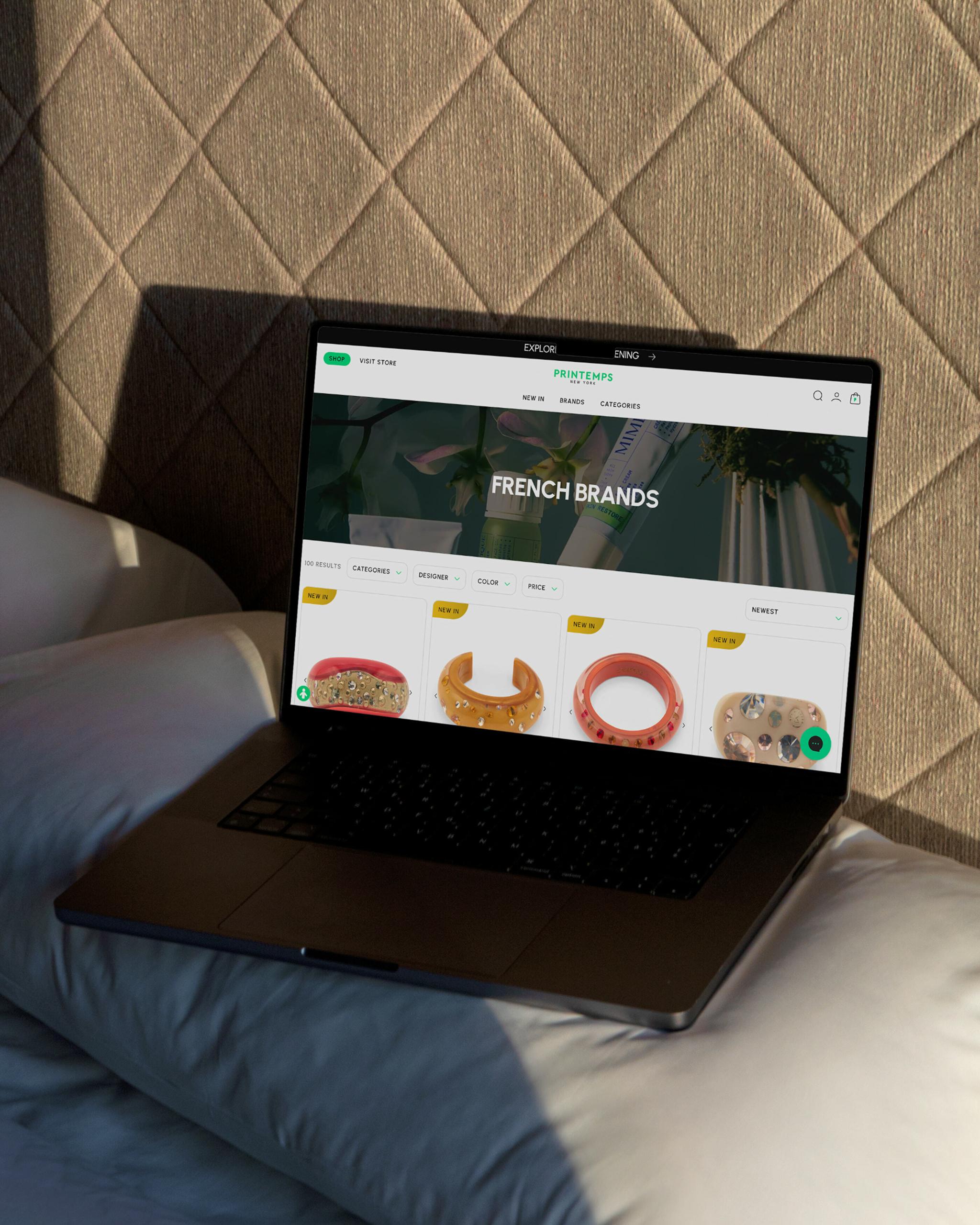 Laptop on a bed showing the Printemps website with French brands and accessories