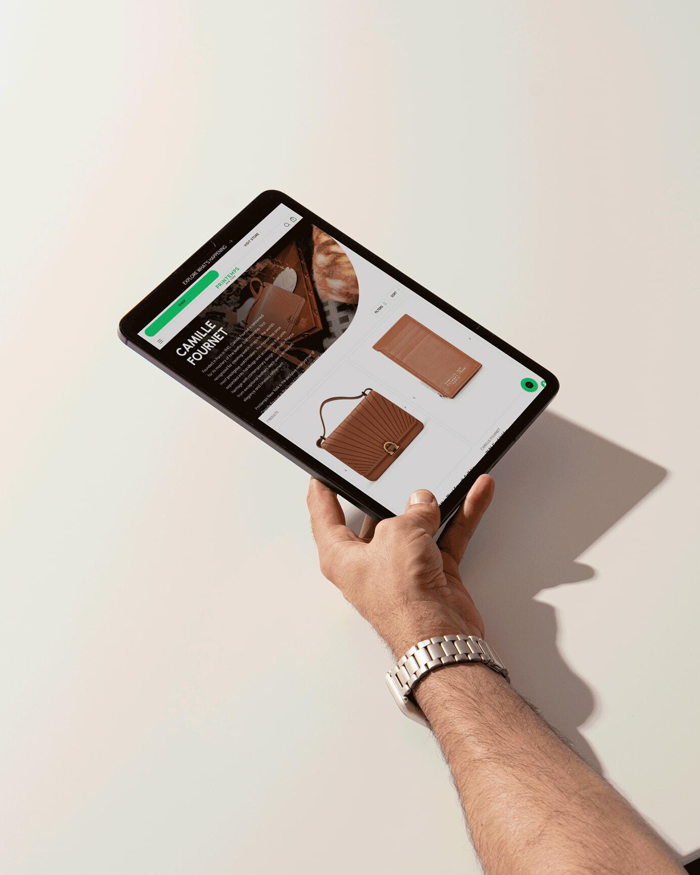 Hand holding a tablet displaying a Printemps product page for brown leather bags