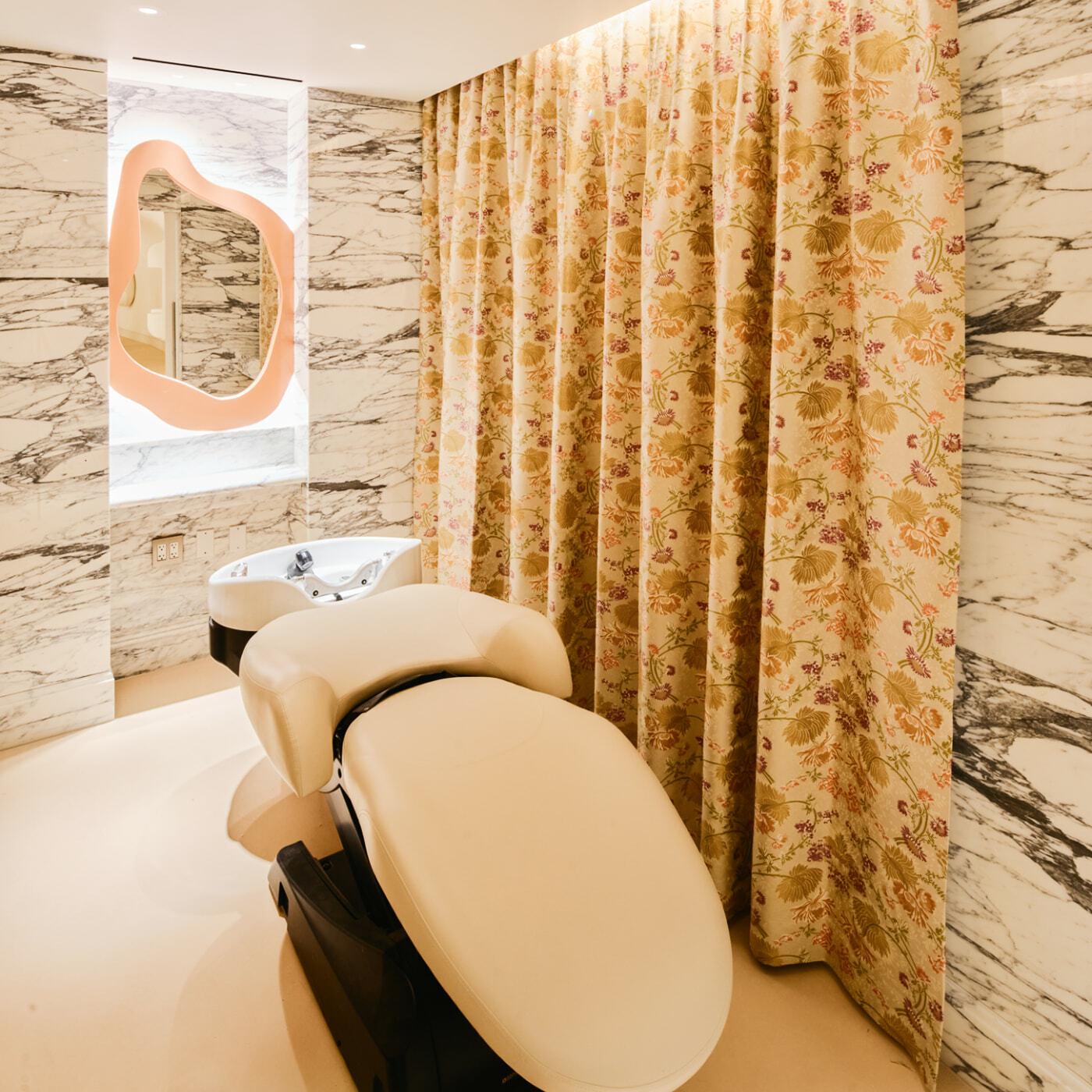 Beauty treatment chair in a marble-walled room with floral curtains and a sink