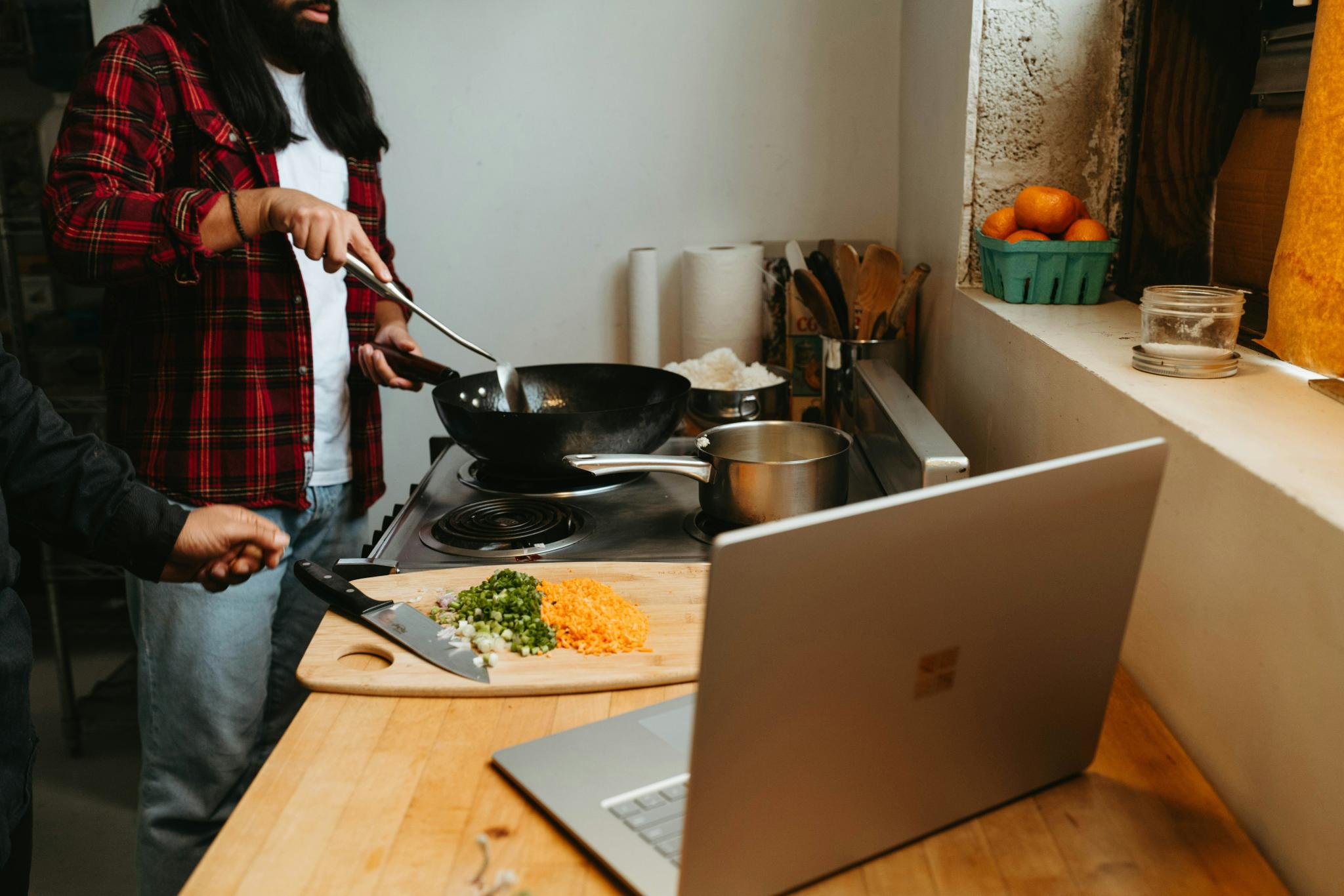 People cooking with a wok beside chopped vegetables and an open laptop