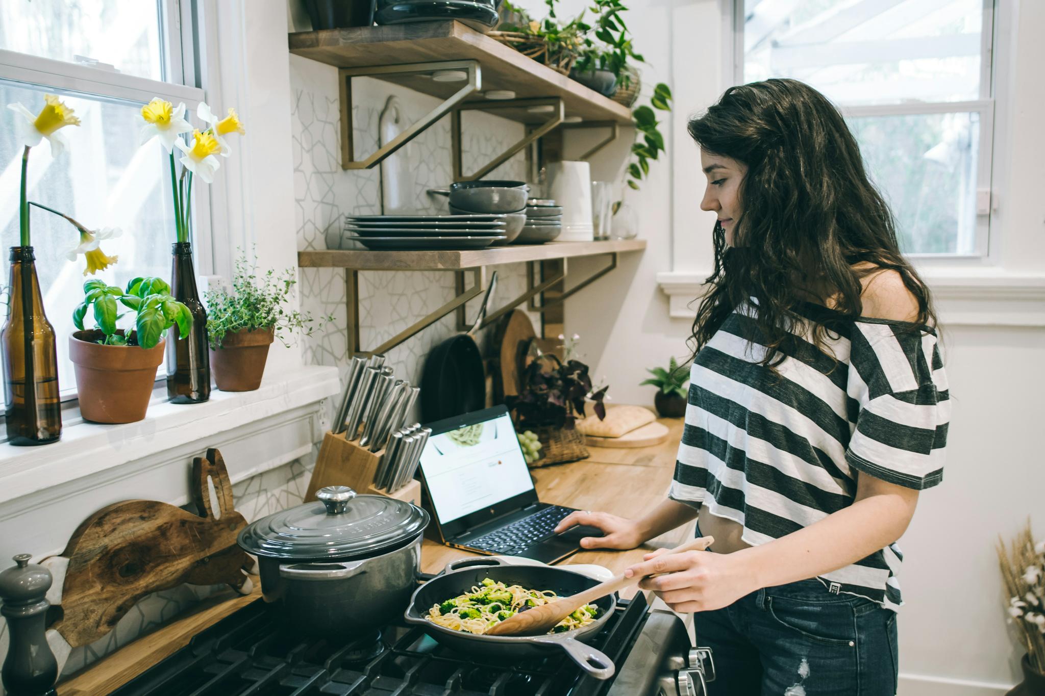Person cooking in a skillet beside an open laptop in a bright kitchen