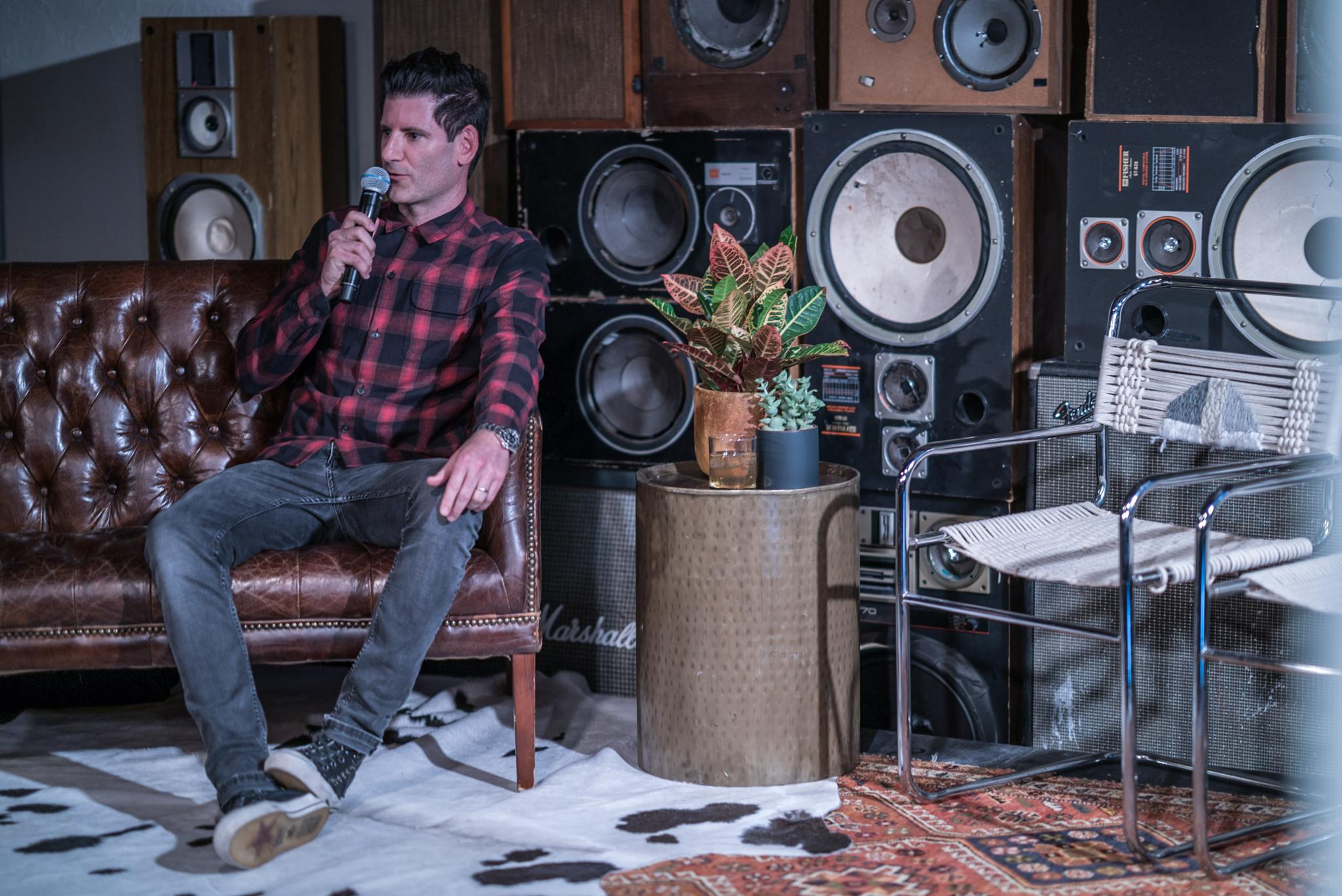Speaker seated with a microphone in a lounge surrounded by speakers