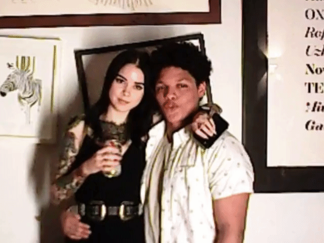 Two people posing indoors beside framed wall art, one holding a drink
