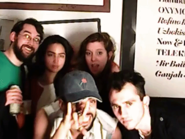 Five people posing closely indoors, some making playful faces, with framed wall art behind them.