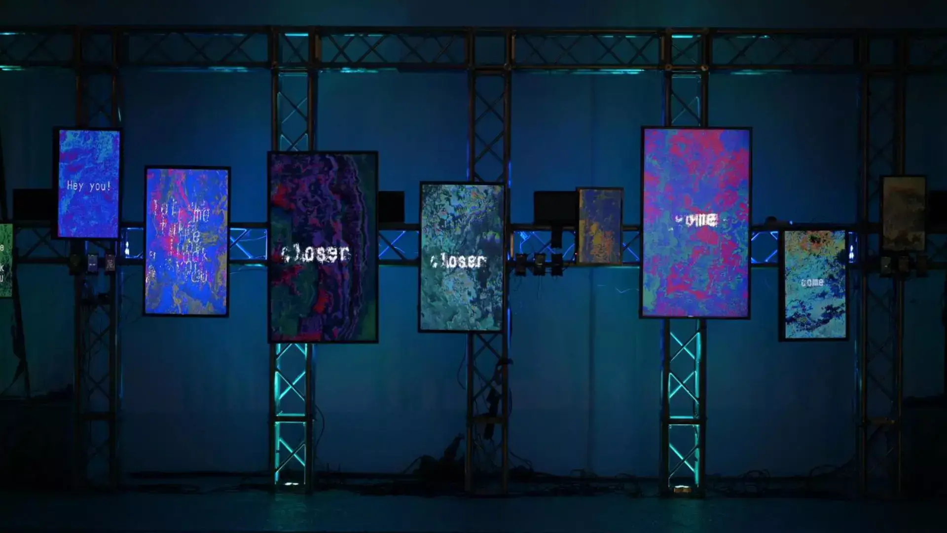 Multiple digital screens on a metal truss display colorful abstract visuals in a dark room.