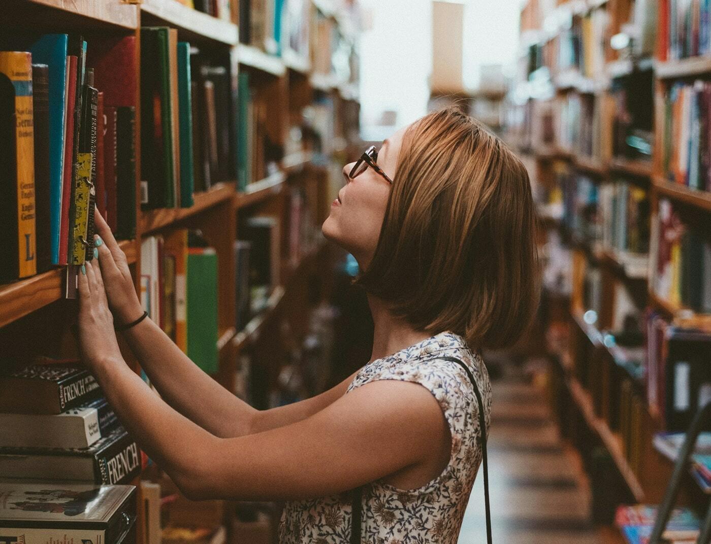 Person browsing books on a library shelf while wearing glasses