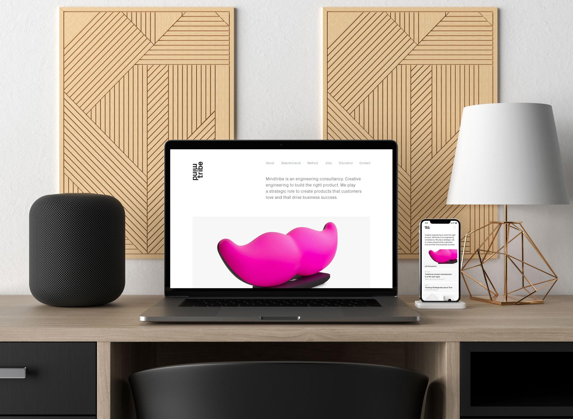 Desk setup with laptop and phone showing a product site, flanked by a speaker and table lamp