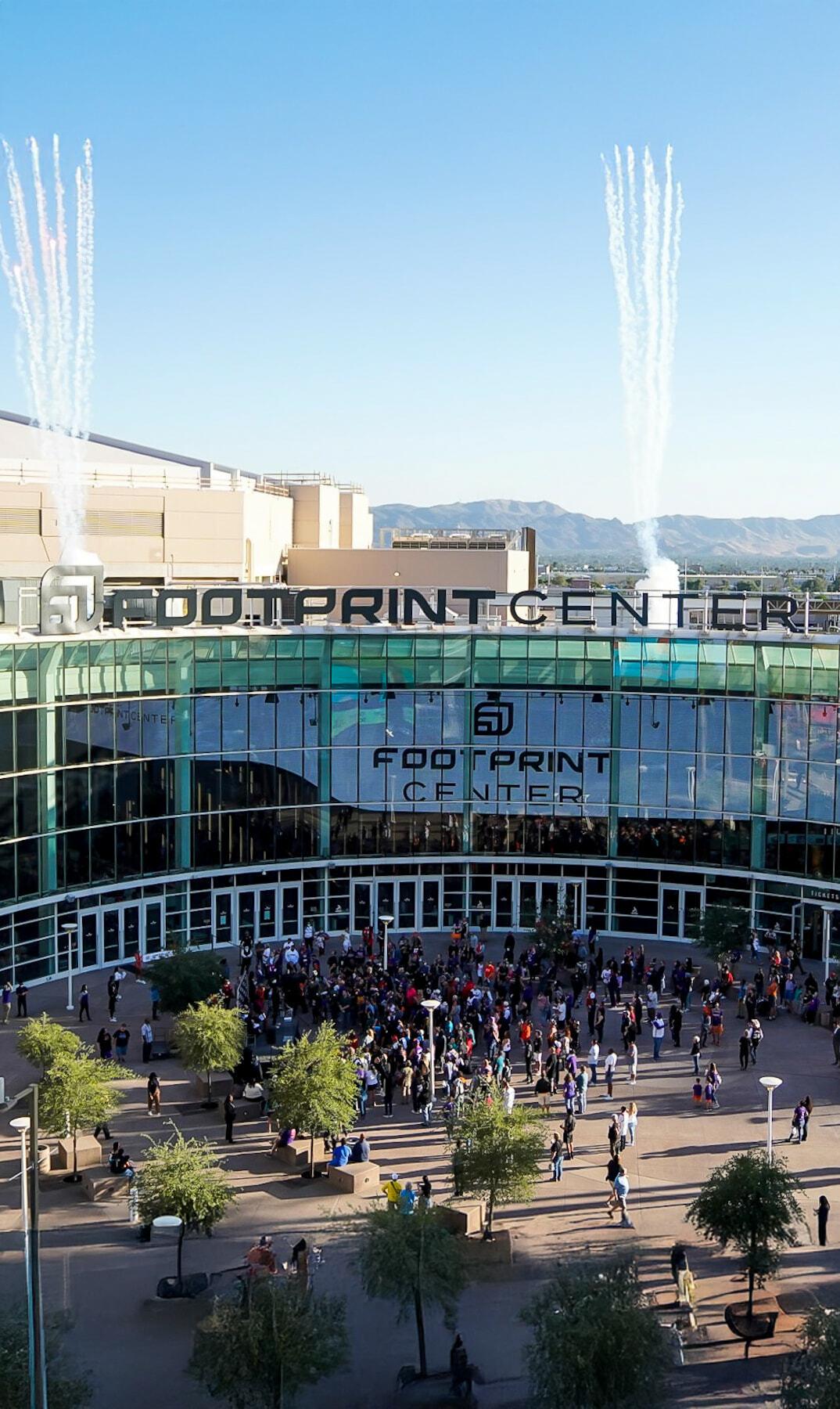 Crowd outside Footprint Center entrance with pyrotechnic smoke trails rising into a clear sky