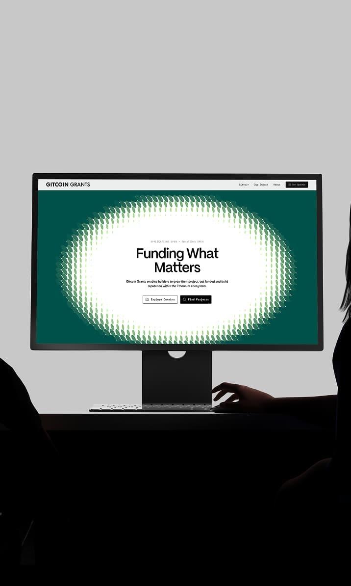 Monitor showing the Gitcoin Grants homepage, with two silhouetted people nearby