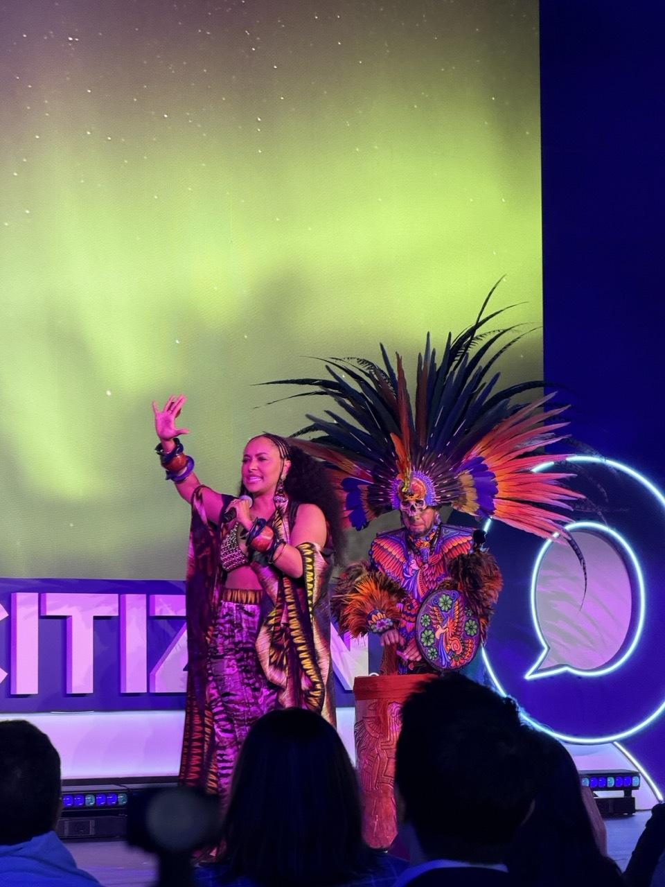 Performers in colorful regalia on a Global Citizen stage under aurora visuals