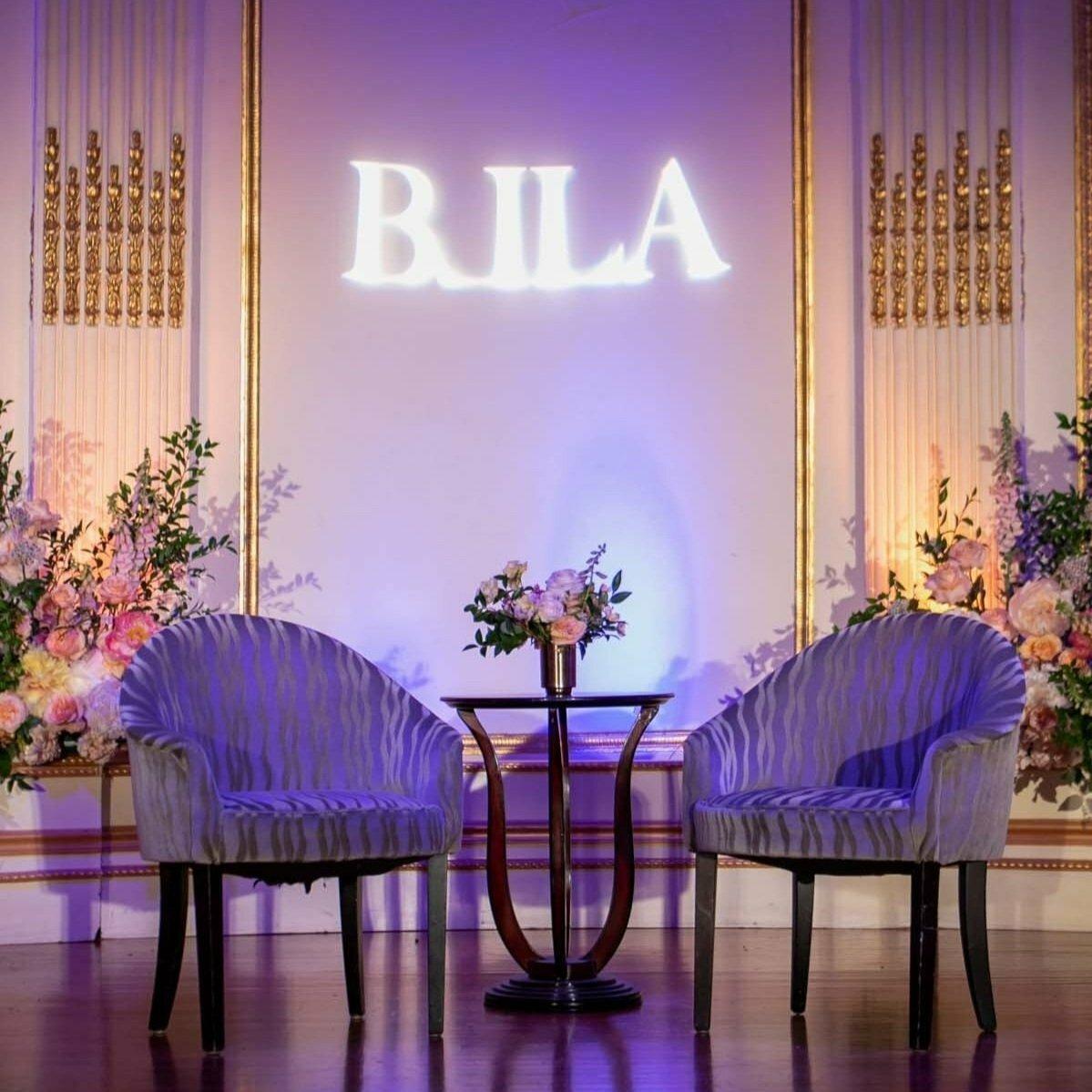 BILA logo projected above two chairs and floral arrangements on a stage