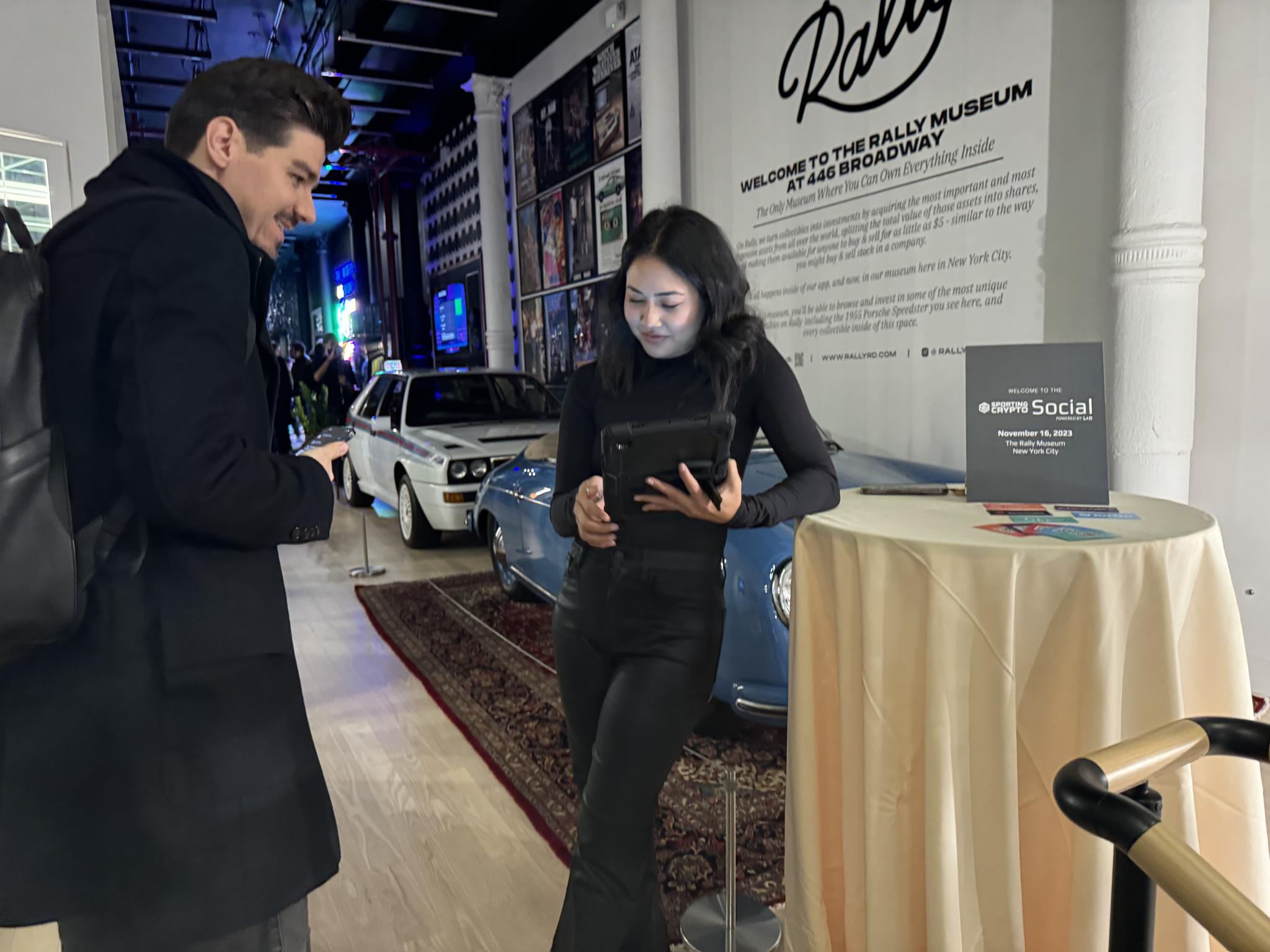 Two attendees using a tablet near cars and a Sporting Crypto Social sign