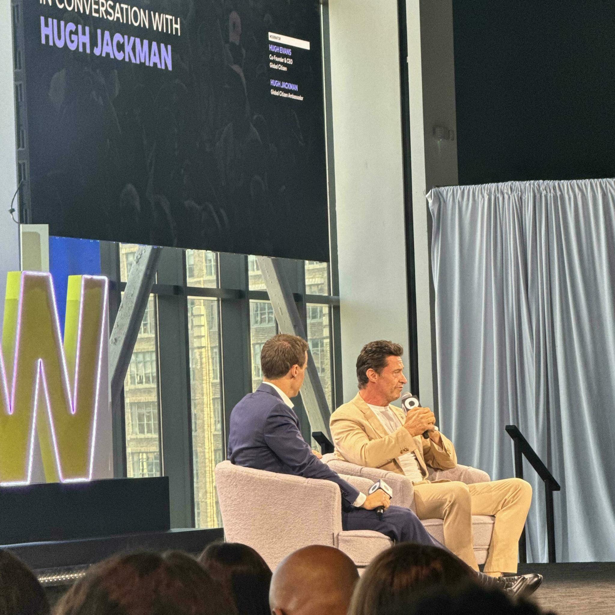 Stage interview with two speakers under "In Conversation with Hugh Jackman" sign
