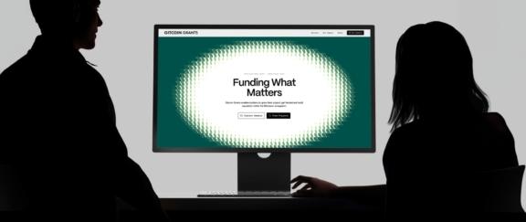Gitcoin Cover Desktop Wide view of two silhouettes facing a monitor with the Gitcoin Grants homepage