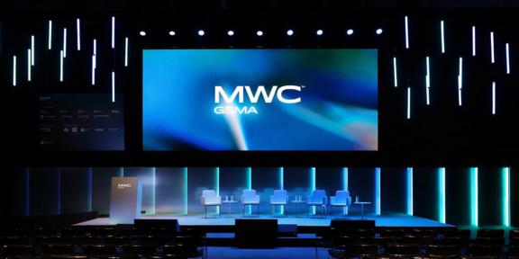 LR exhibiting at Mobile World Congress Barcelona 2026 MWC GSMA conference stage with large screen, podium, chairs, and blue vertical lights