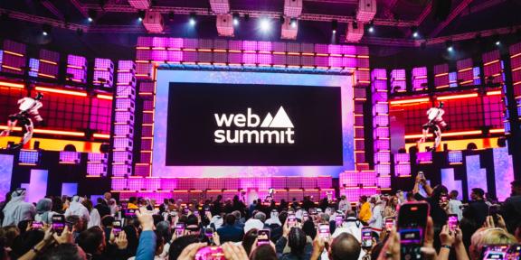 Web Summit Qatar 2026 Crowd recording a brightly lit Web Summit stage with large screen and pink lighting.