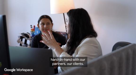 Two coworkers discuss at a desk with Google Workspace caption on screen