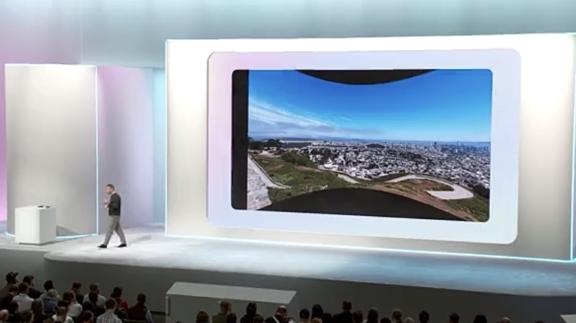 Presenter on stage beside a large screen showing a city landscape