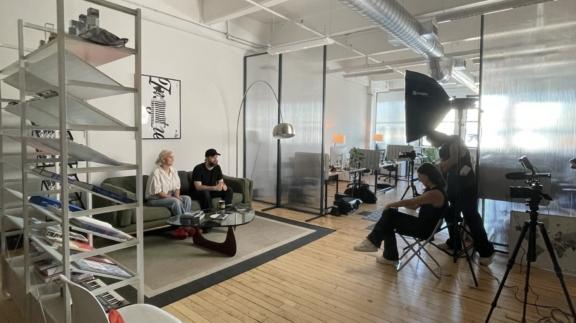 Film crew interviewing two people in a bright studio with cameras, lights, and shelving.