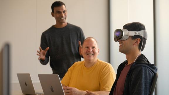Three coworkers in a meeting, one wearing a mixed reality headset beside open laptops