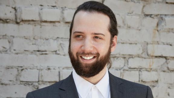 Smiling bearded man in a suit and white shirt against a white brick wall