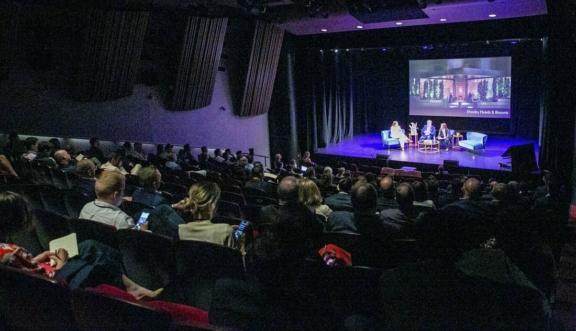 Audience watching a panel discussion on a theater stage with a large presentation screen