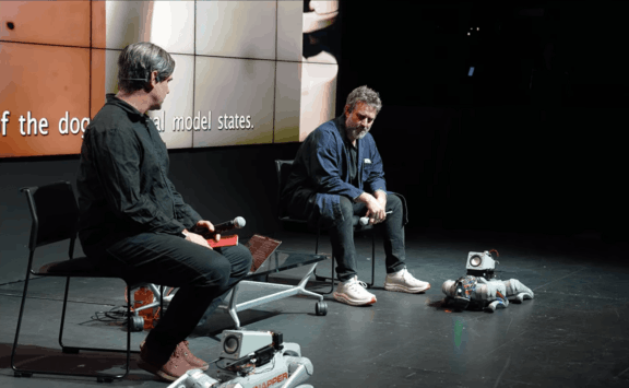 SIGGRAPH session showing two speakers seated near companion robot dogs on a stage.