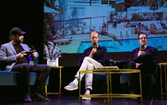 Three panelists seated on stage with a pool resort image projected behind them