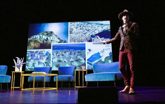 Presenter on stage beside blue chairs and a screen showing resort images