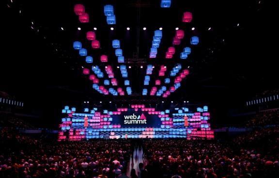 Large Web Summit auditorium with colorful suspended lights and audience