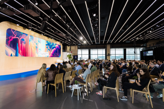 Audience seated in a modern conference hall facing a large colorful presentation screen