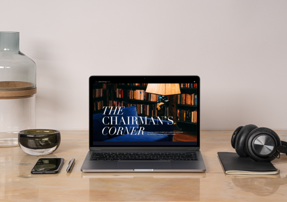 Laptop on a desk showing The Chairman's Corner, with headphones and office items