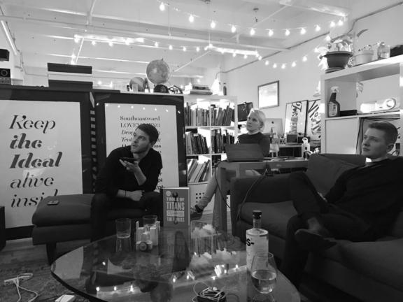 Three people in a cozy office lounge with bookshelves, posters, string lights, and a glass coffee table