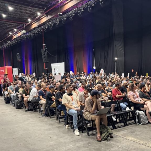 Large audience seated in a conference hall with stage lighting and black curtains