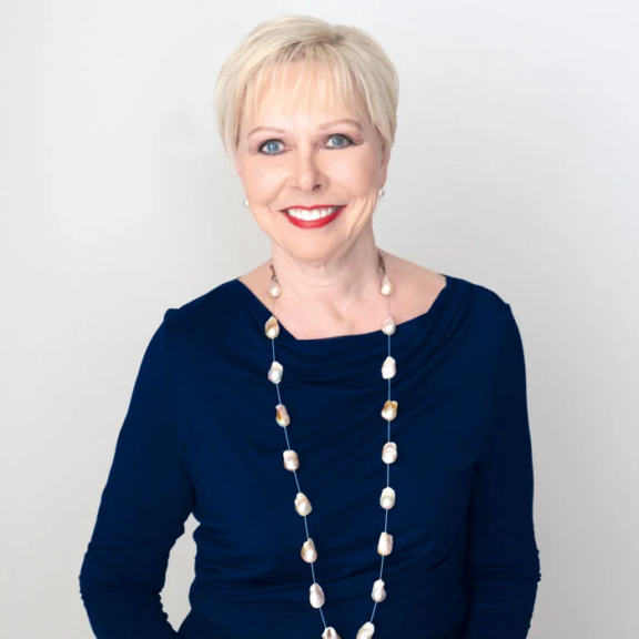 Marilumarshall Smiling woman with short blonde hair wearing a navy top and pearl necklace on a light background