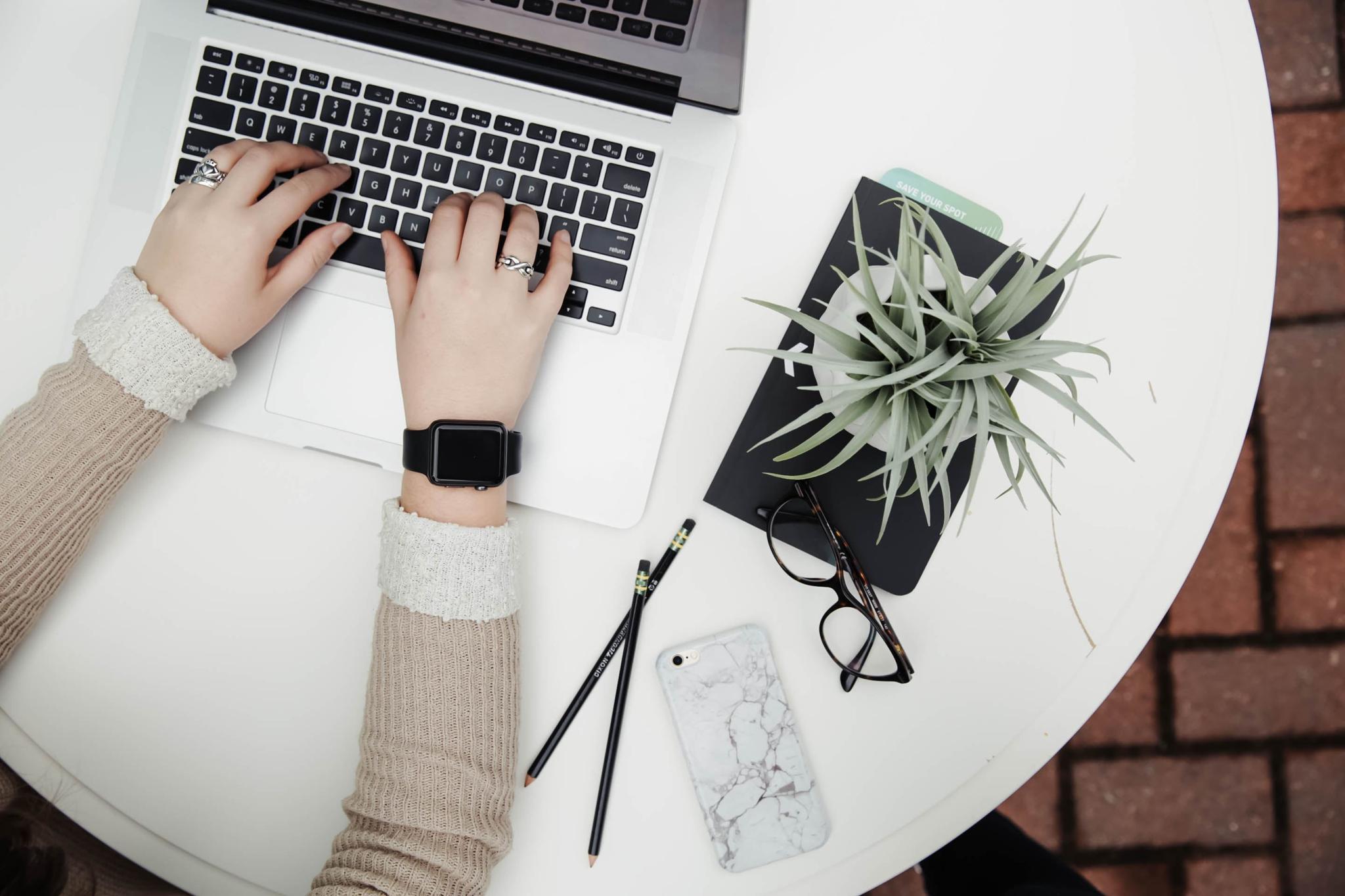 Hands typing on a laptop at a round desk with plant and notebook