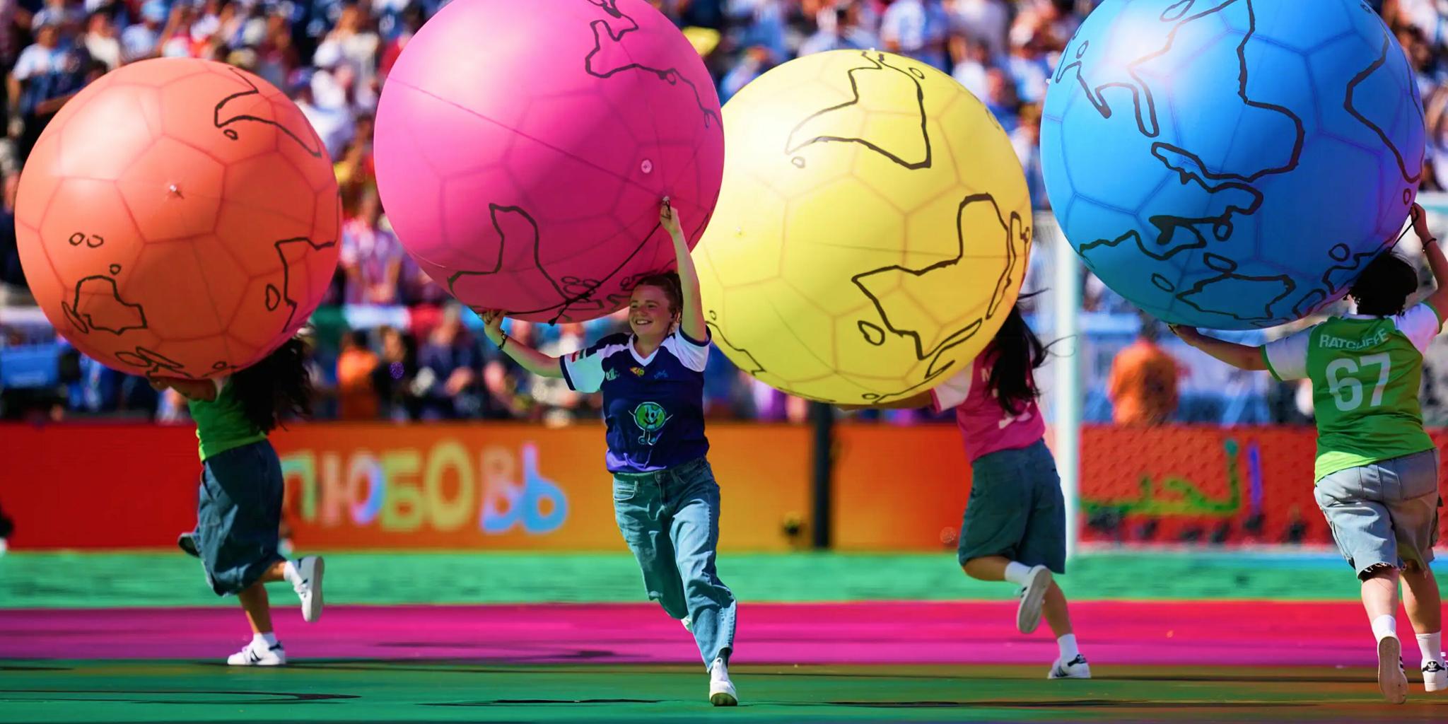 Performers carry oversized colorful soccer balls across a stadium field.