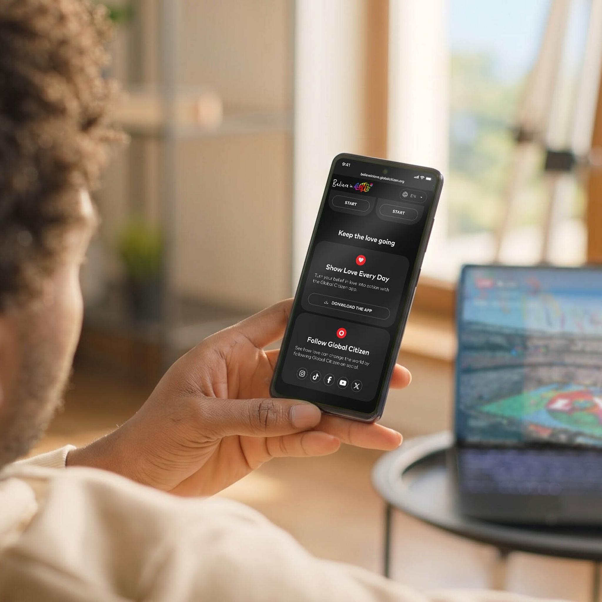 Person holding a phone with a dark campaign app screen beside a laptop.