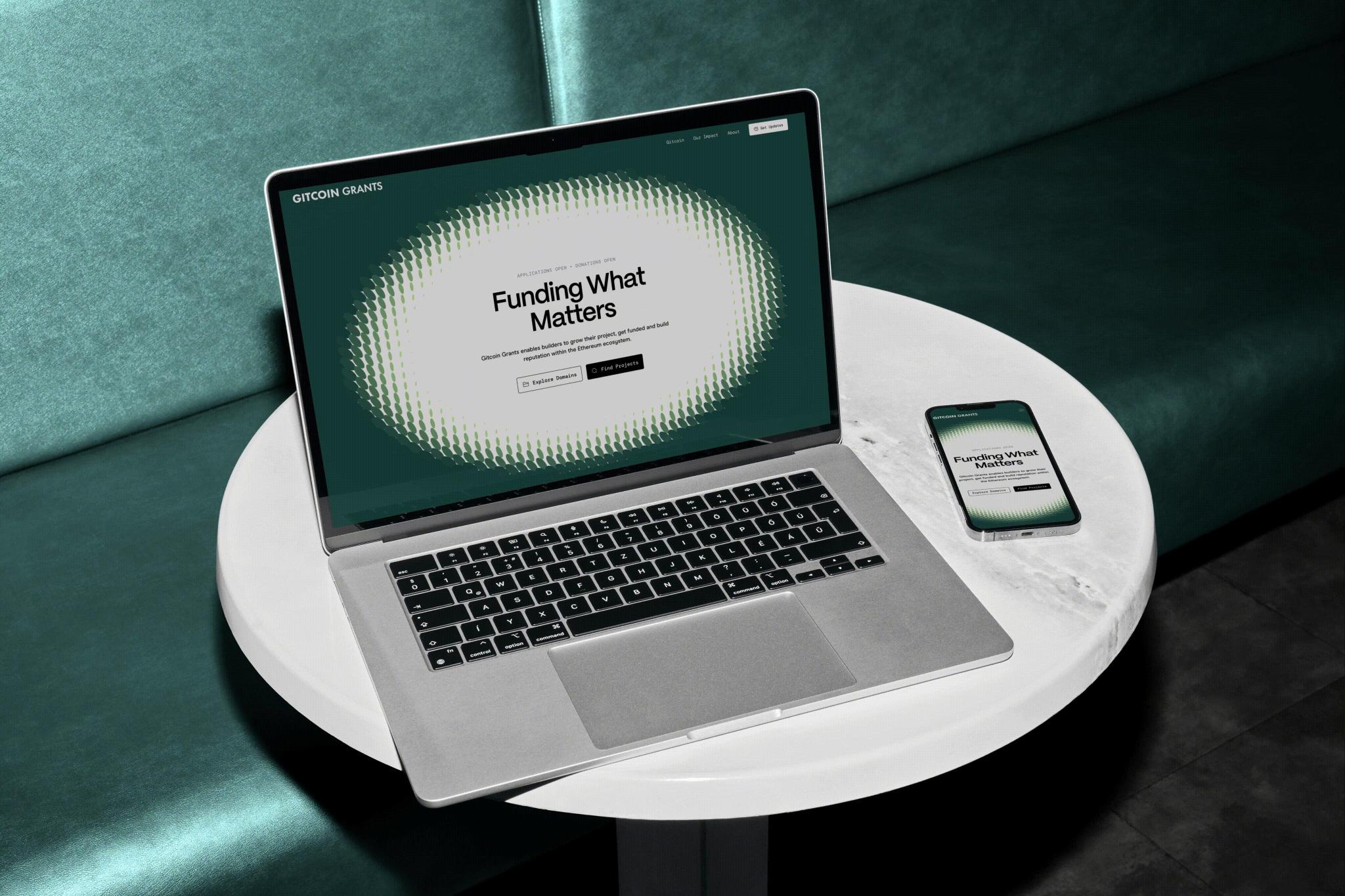 Laptop and phone on a white table display the Gitcoin Grants homepage.
