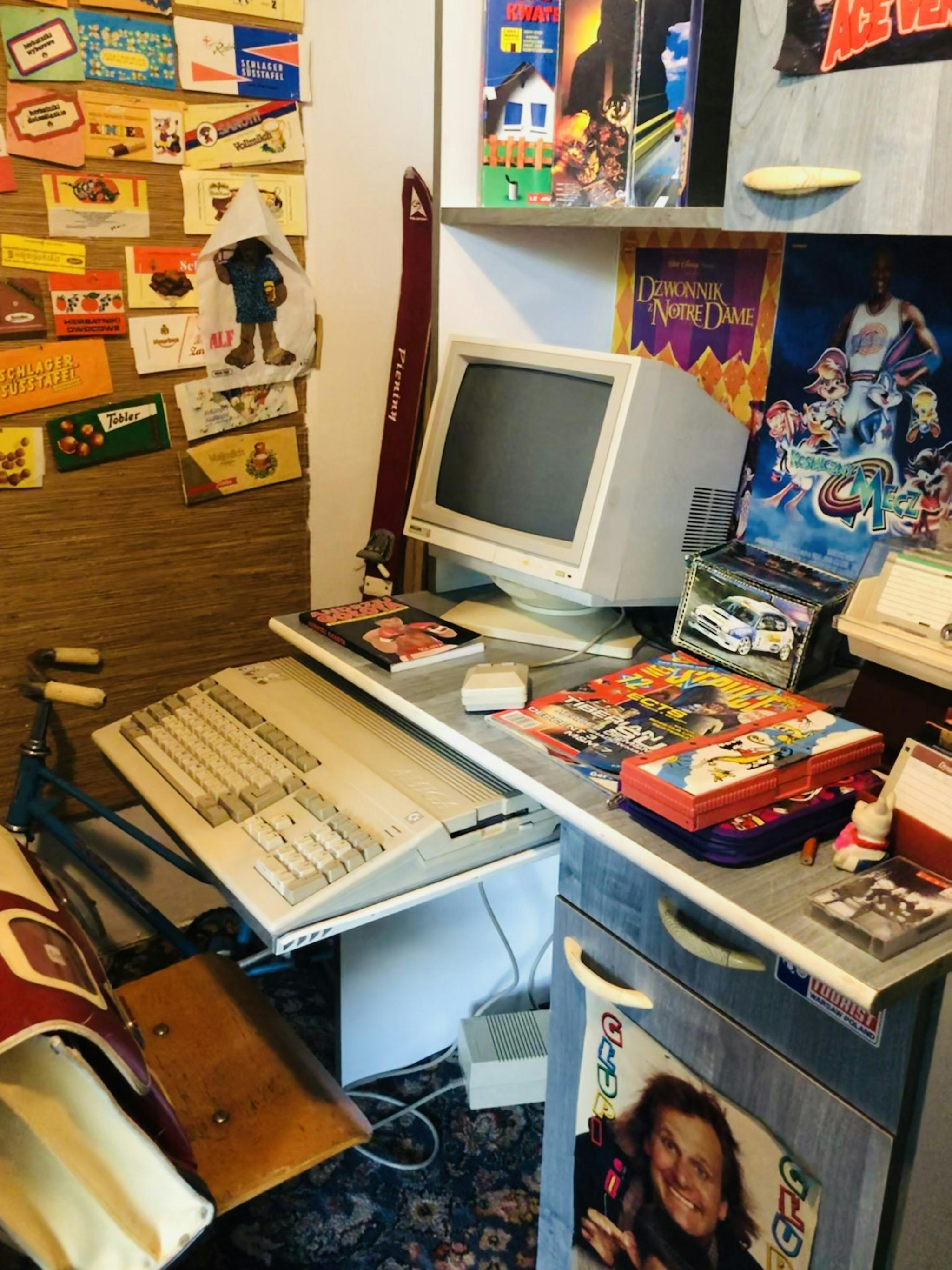 Cluttered retro desk with a CRT computer, keyboard, game boxes and pinned posters