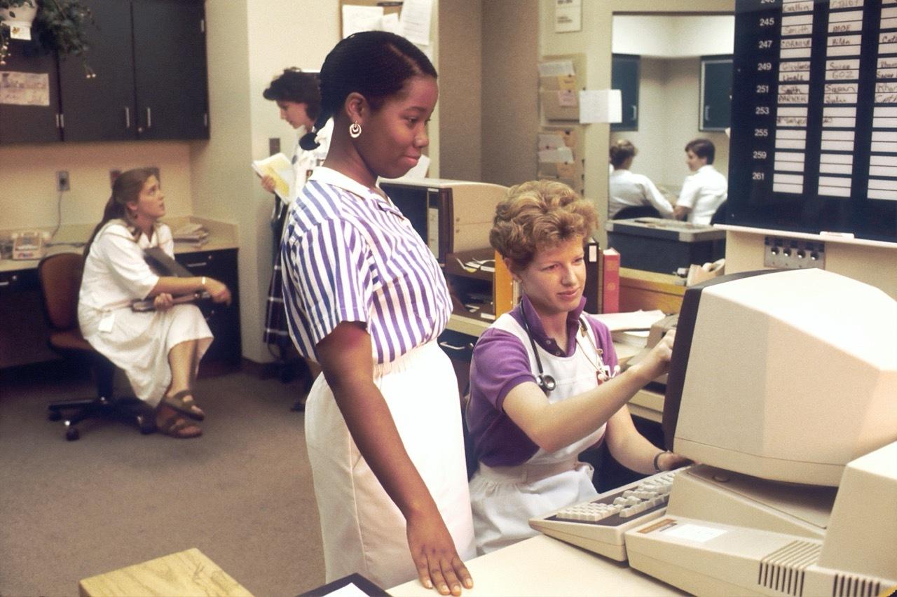 Nurses working at a beige desktop terminal in a 1980s hospital office