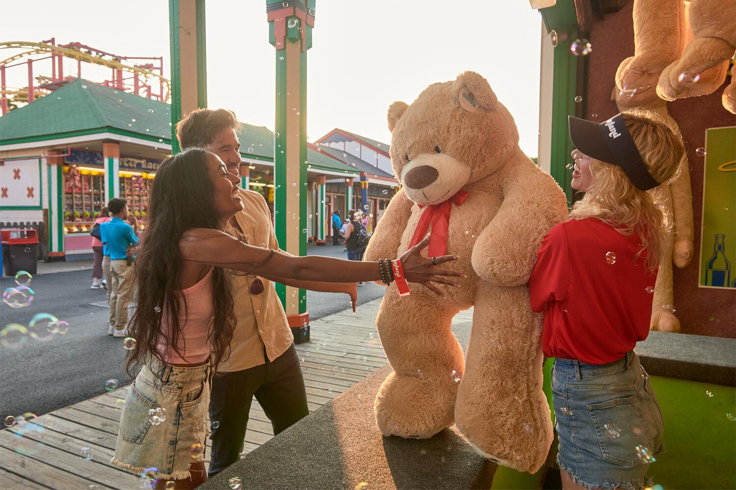 People reaching toward a giant teddy bear prize at an amusement park game