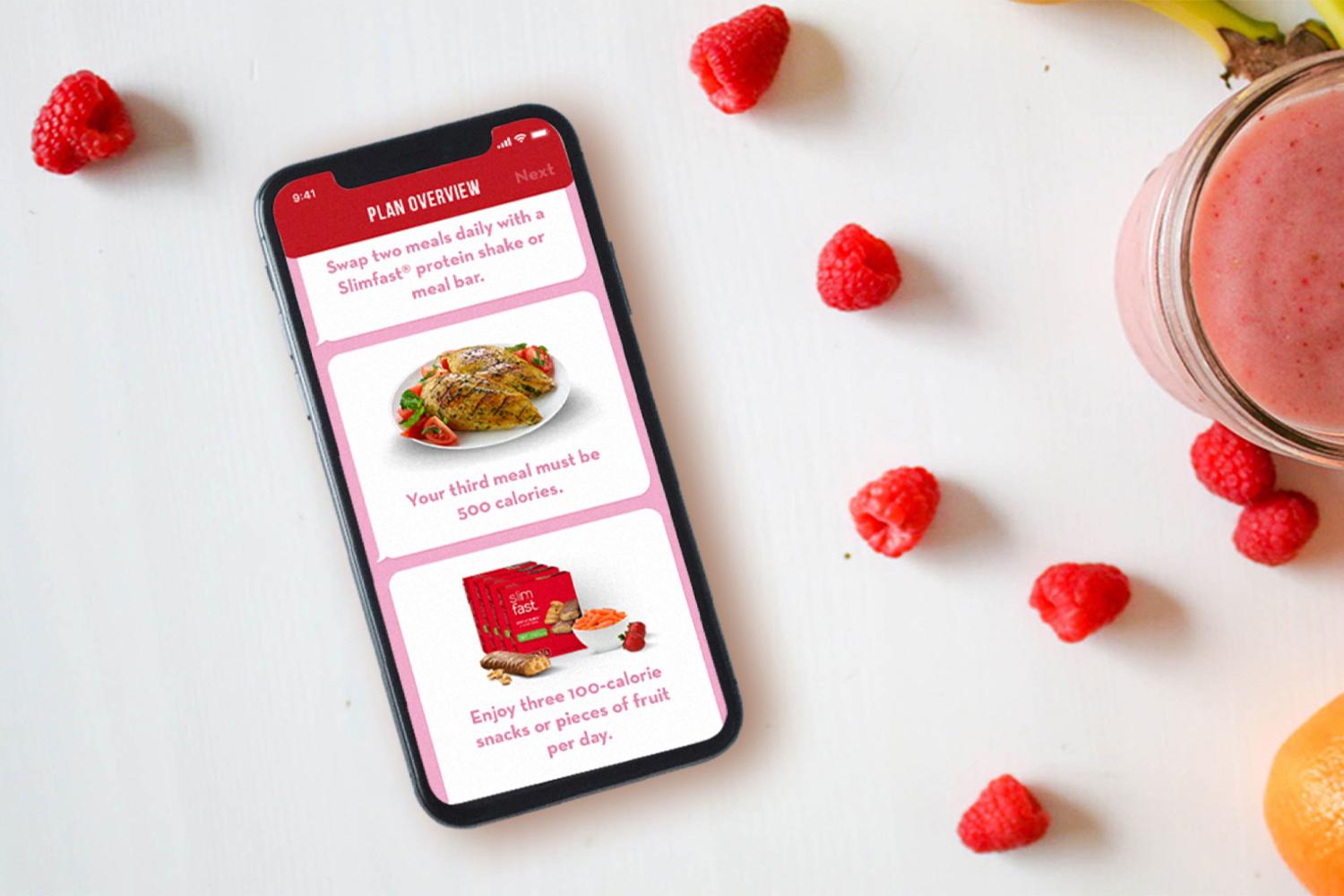 Smartphone showing a diet plan app beside raspberries, a smoothie, banana, and orange